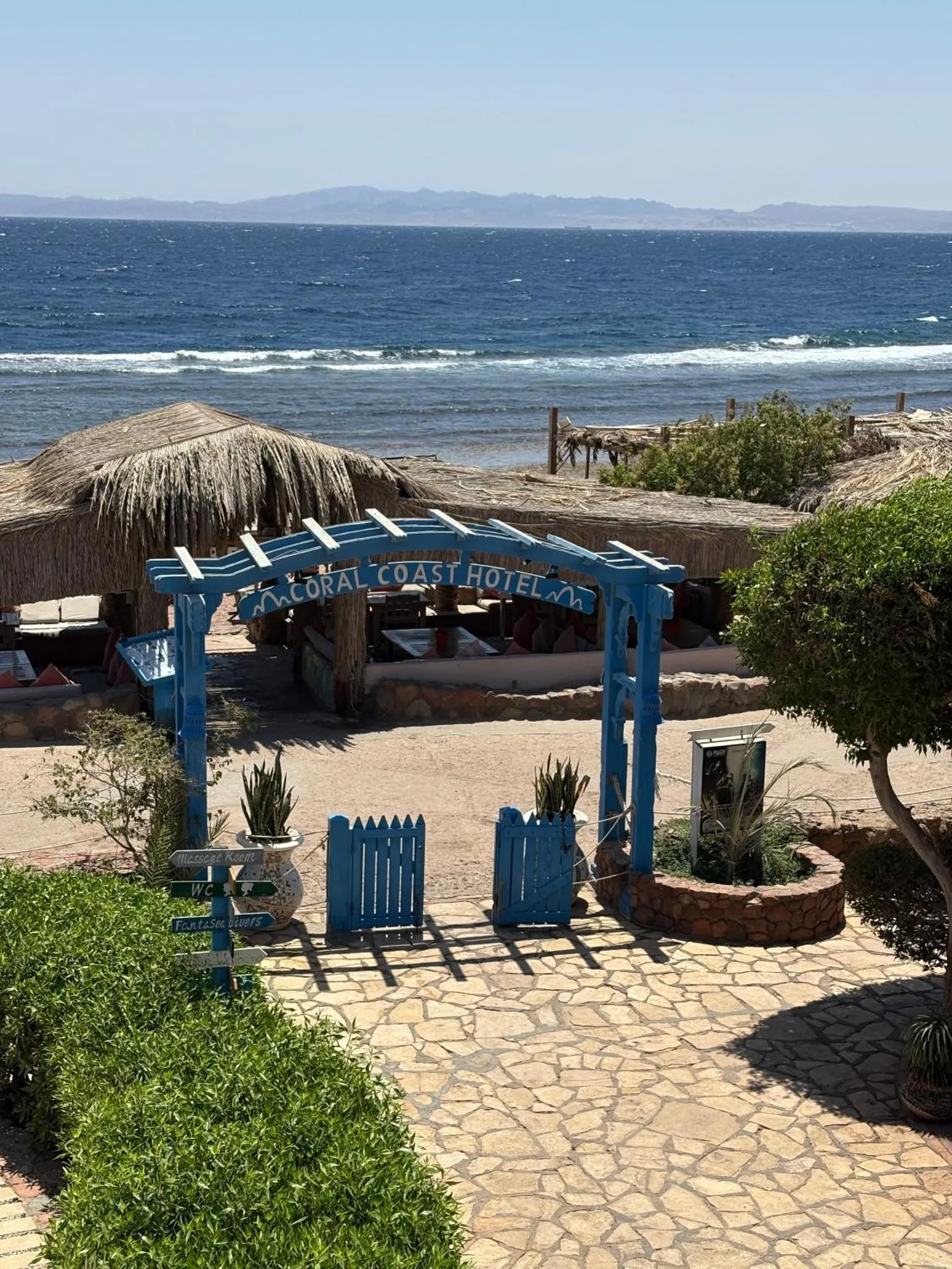 Vue d'une entrée ombragée avec végétation et mobilier bleu face à la mer.