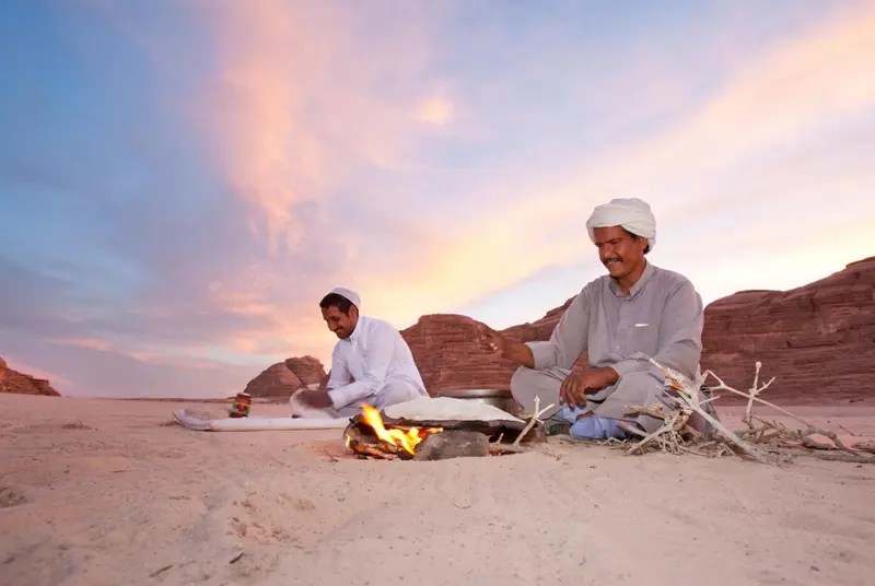 Deux hommes préparent un repas sur un feu de camp dans un paysage désertique au coucher du soleil.