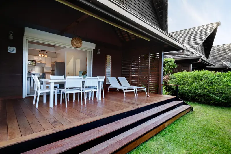 Terrasse en bois avec table à manger et chaises blanches, donnant sur une cuisine ouverte.
