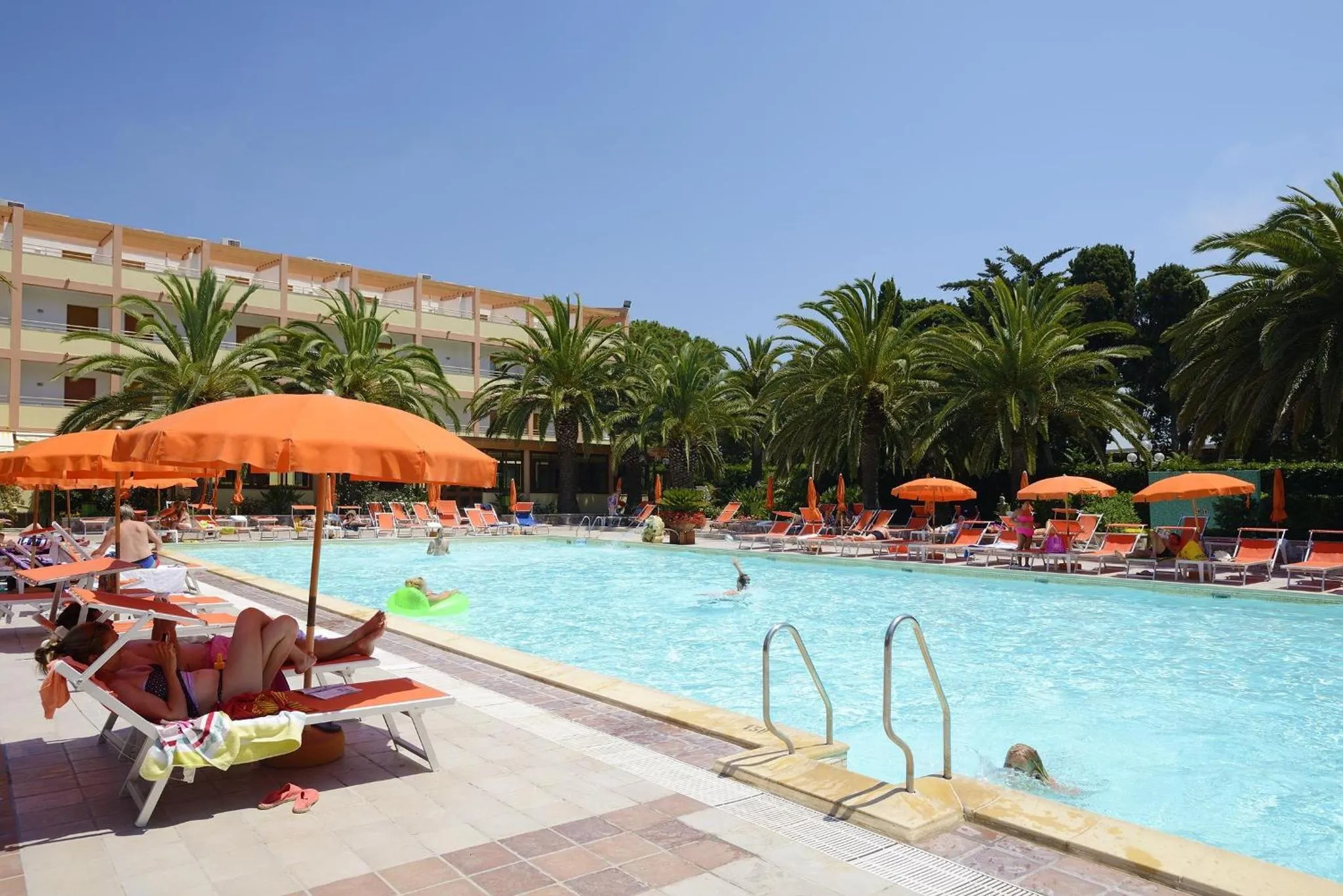 Transats orange alignés au bord d'une piscine ensoleillée, avec un bâtiment en arrière-plan.