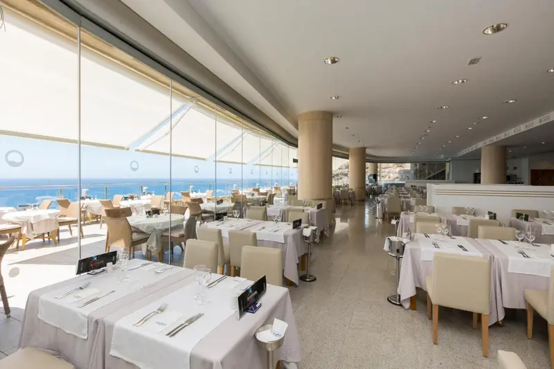 Une salle de restaurant lumineuse avec de grandes baies vitrées donnant sur la mer.