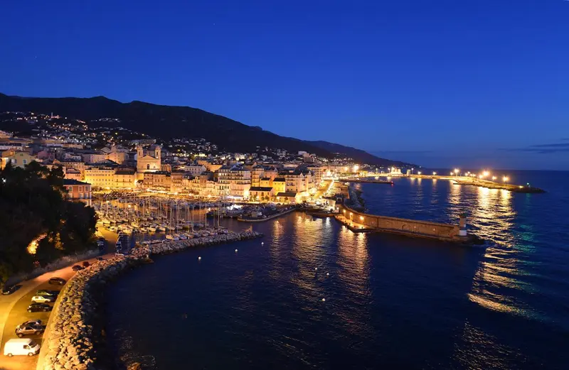 Vue nocturne d'une ville côtière illuminée avec un port de plaisance.