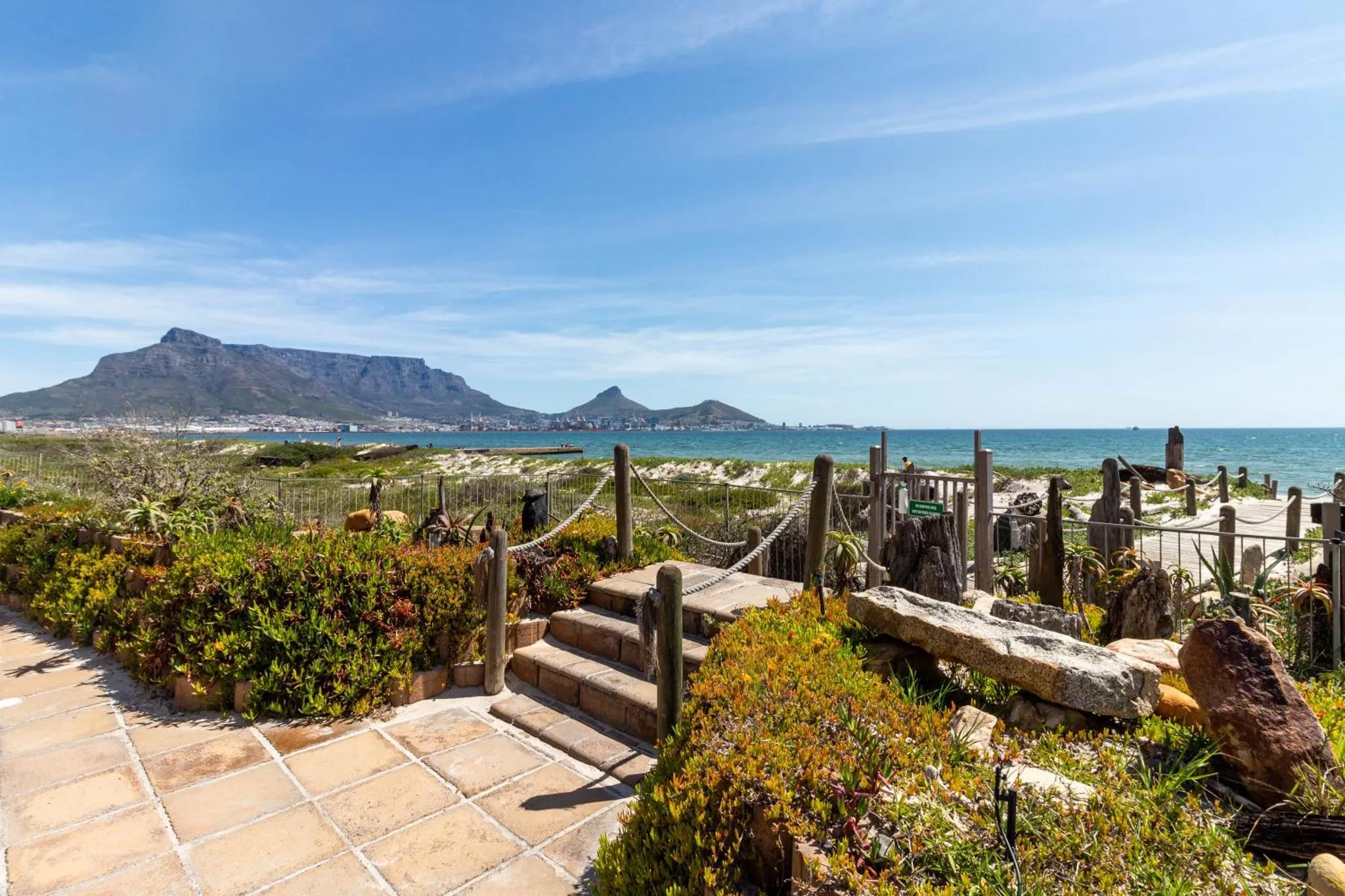 Un jardin fleuri mène à une plage de sable fin avec une vue sur la mer et des montagnes au loin.
