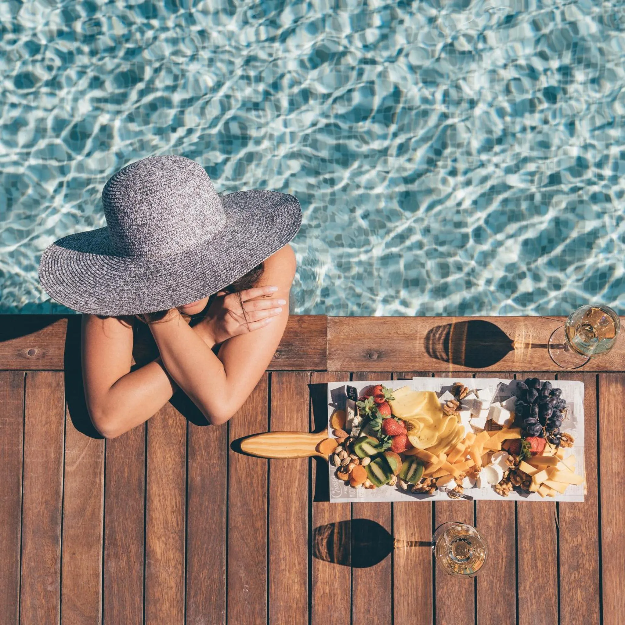 Une personne vue de dos se détend au bord d'une piscine avec un chapeau et une assiette de fruits.