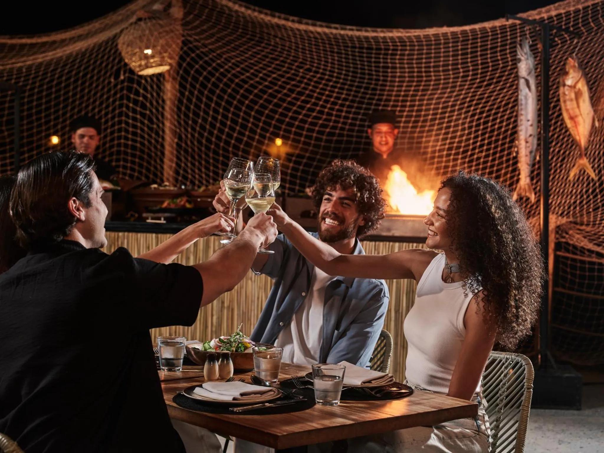 Des convives trinquent avec des verres de vin autour d'une table éclairée par un feu de joie.