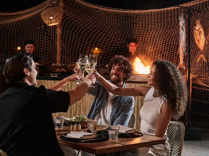 Des convives trinquent avec des verres de vin autour d'une table éclairée par un feu de joie.