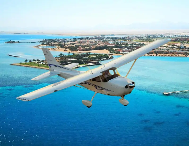 Un avion léger survole une côte turquoise avec des îles et une ville en arrière-plan.