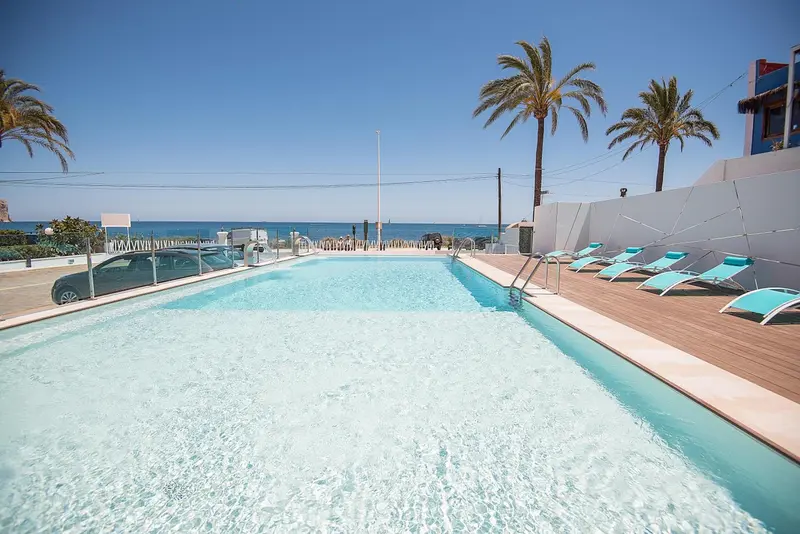 Piscine rectangulaire avec transats bleus et palmiers en arrière-plan.