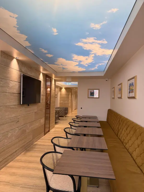 Intérieur lumineux avec des tables et des banquettes le long d'un mur texturé, sous un plafond décoré d'un ciel nuageux.