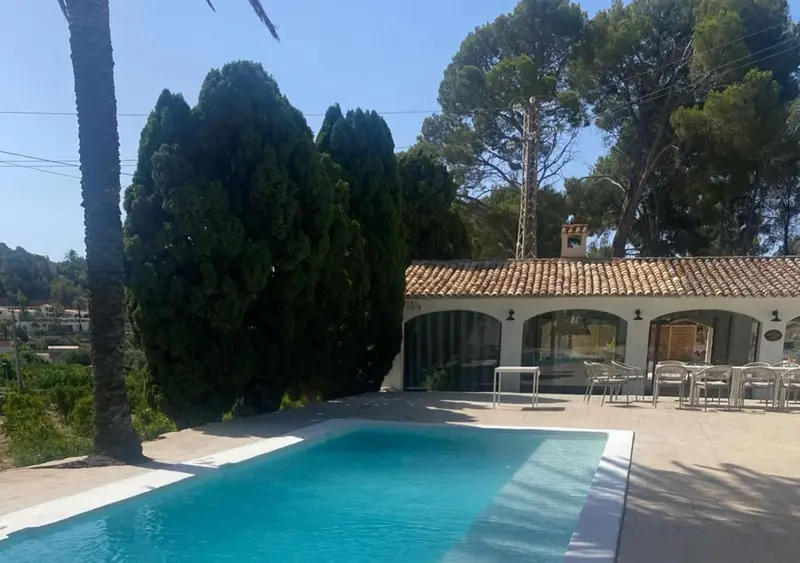 Une piscine turquoise bordée d'une terrasse ensoleillée devant un bâtiment blanc.