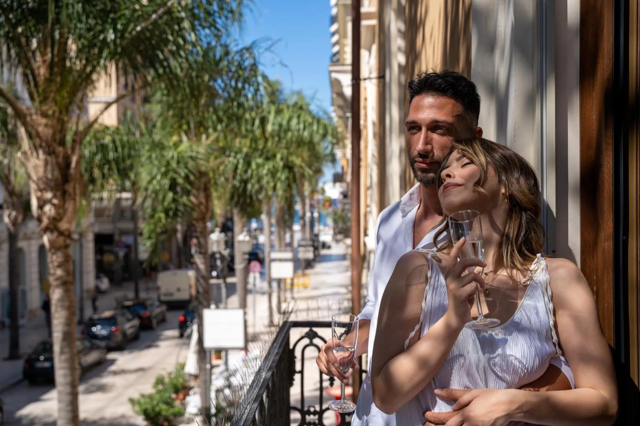 Un couple se tient sur un balcon ensoleillé, levant des verres à champagne.