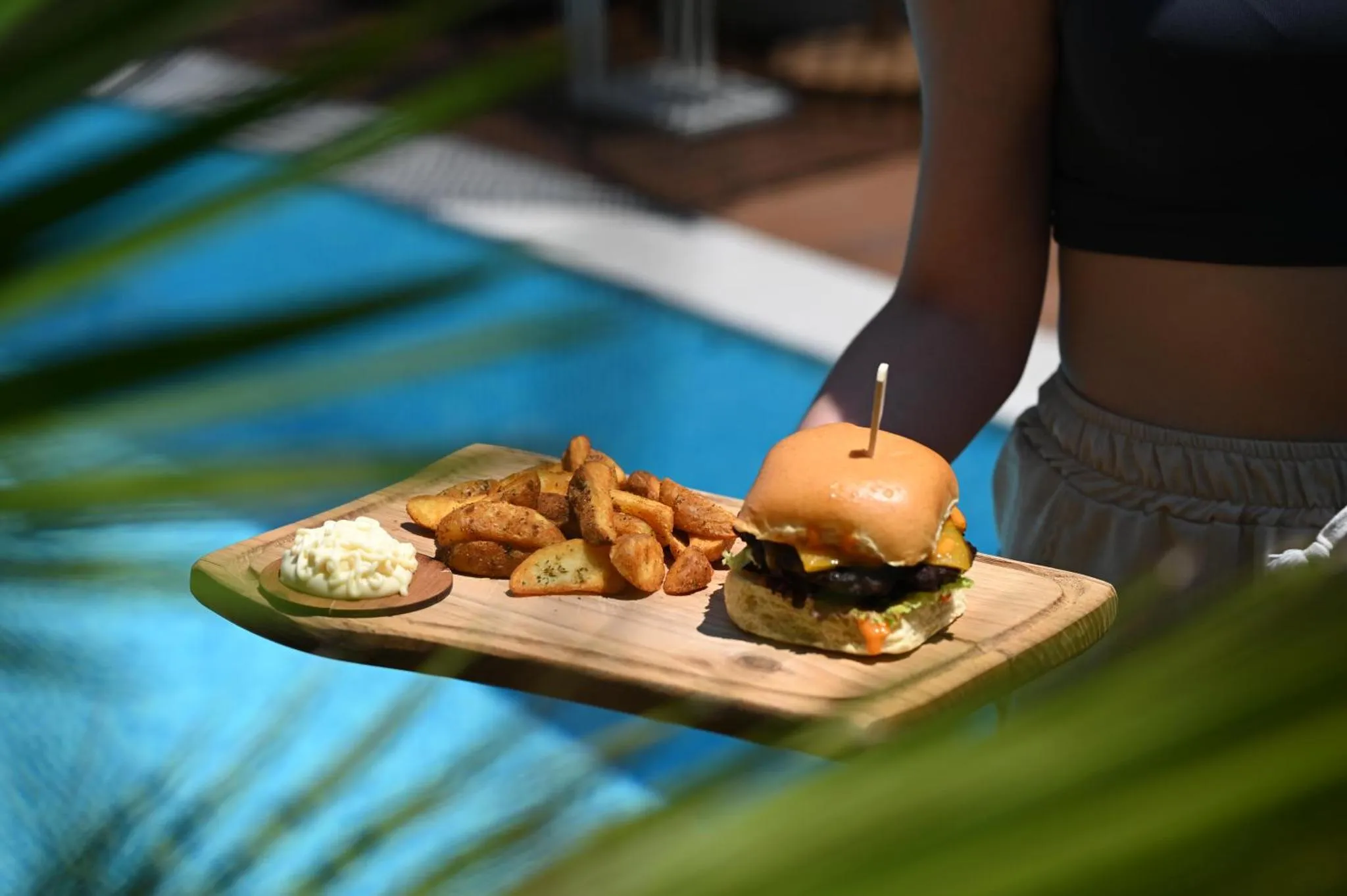 Une personne porte un plateau avec un hamburger et des frites devant une piscine.