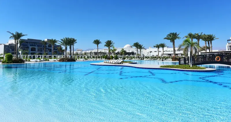 Une grande piscine turquoise avec des palmiers et des bâtiments blancs en arrière-plan.