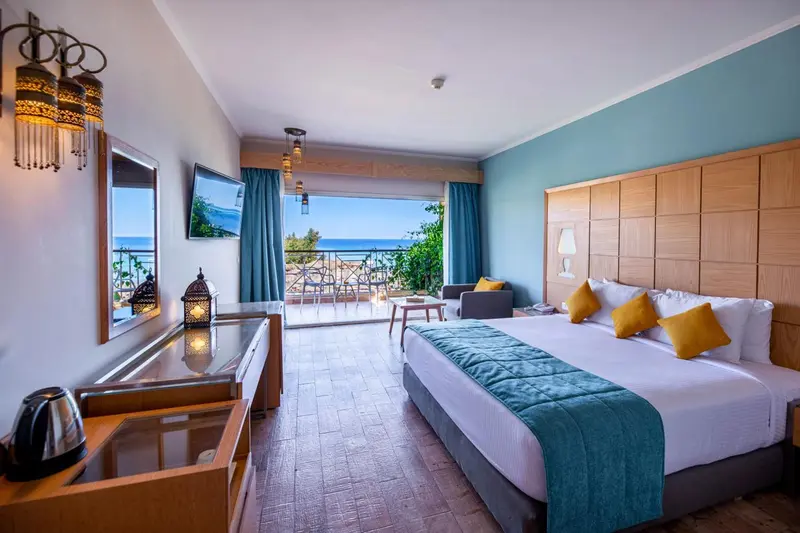 Chambre lumineuse avec un grand lit et une vue sur la mer depuis un balcon.