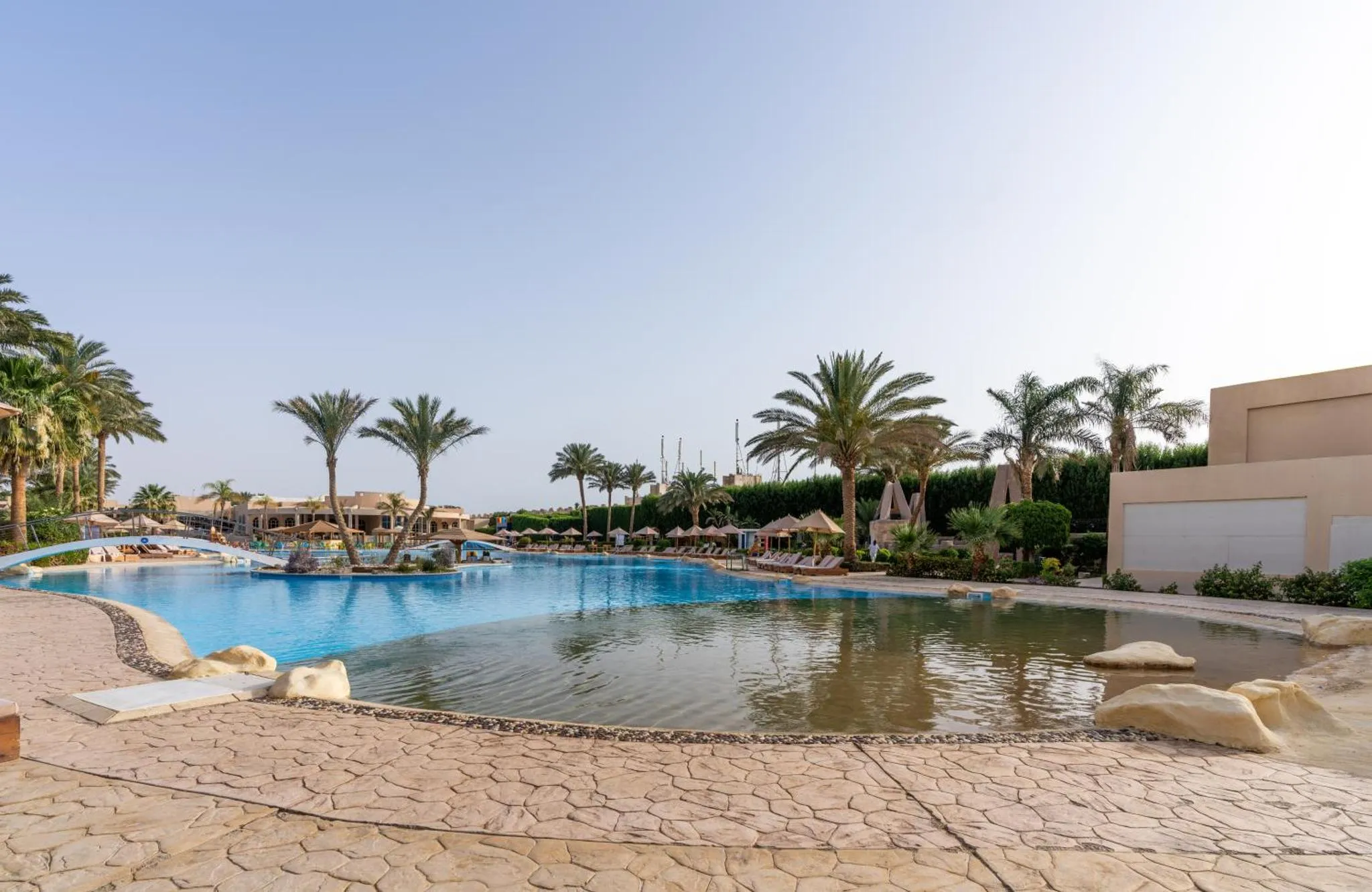 Une piscine d'hôtel avec des transats et des palmiers sous un ciel ensoleillé.