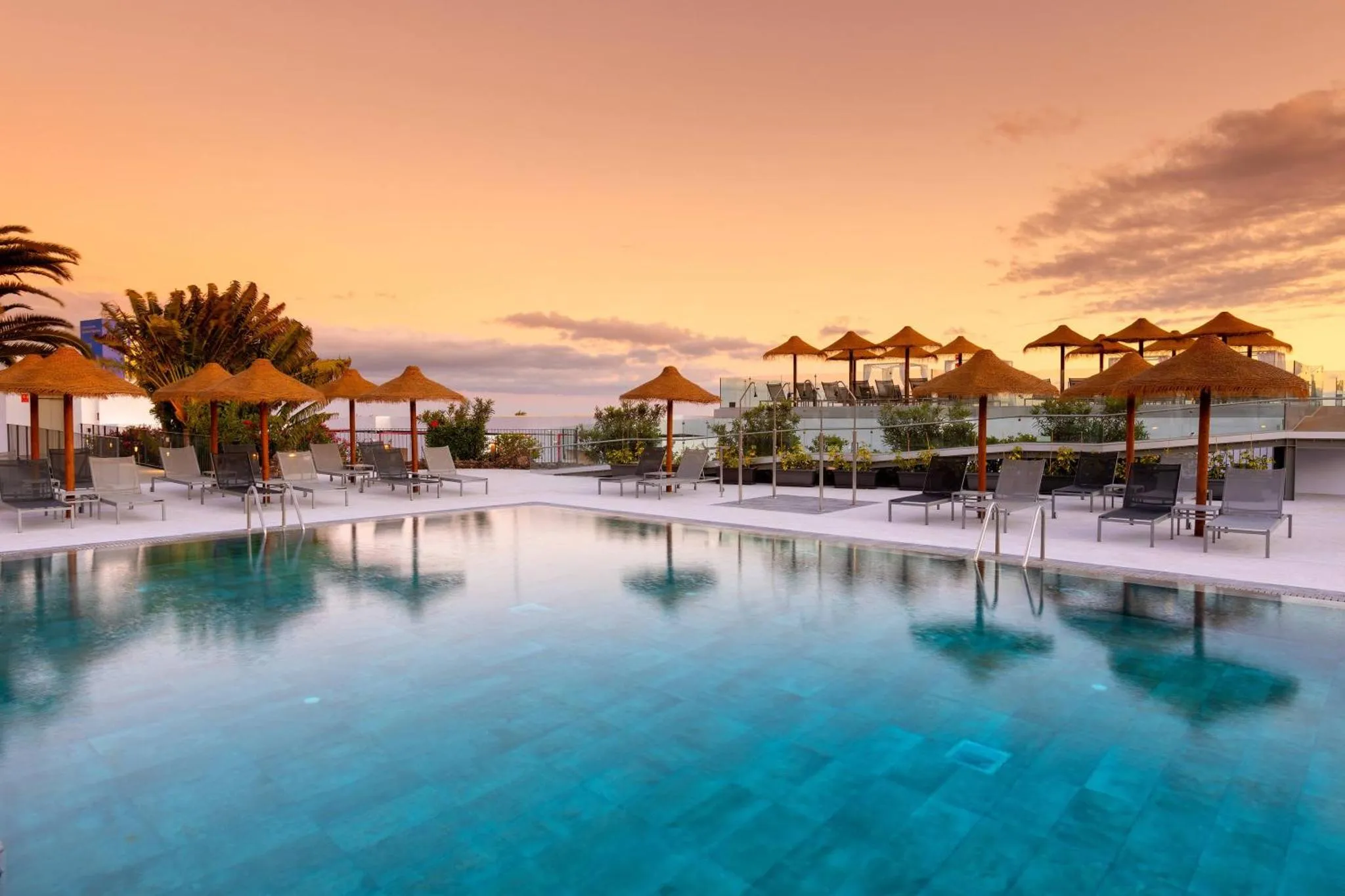 Terrasse avec transats et parasols au bord d'une piscine au coucher du soleil.