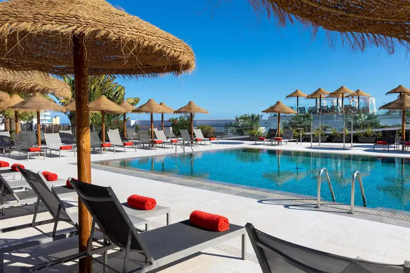 Transats rouges et parasols en chaume bordent une piscine rectangulaire.