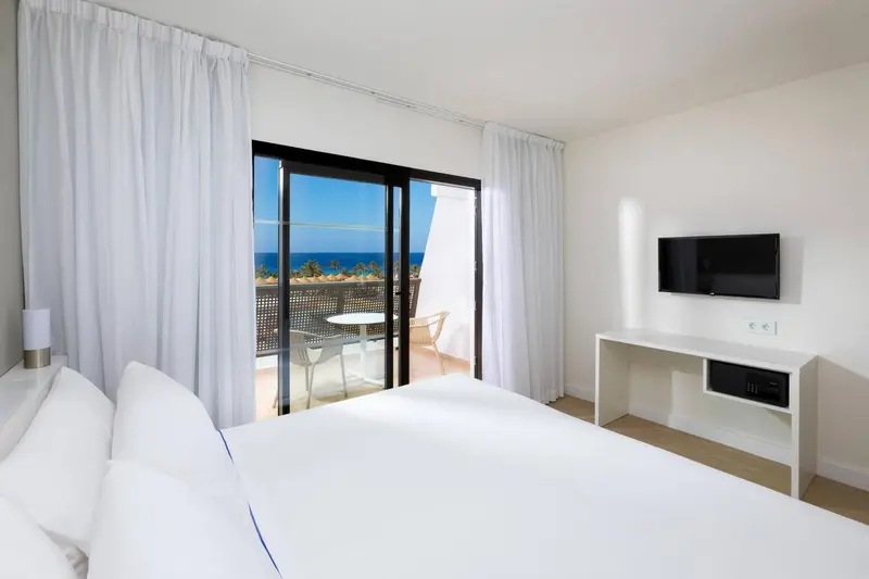 Chambre lumineuse avec un lit blanc et une vue sur la mer depuis un balcon.