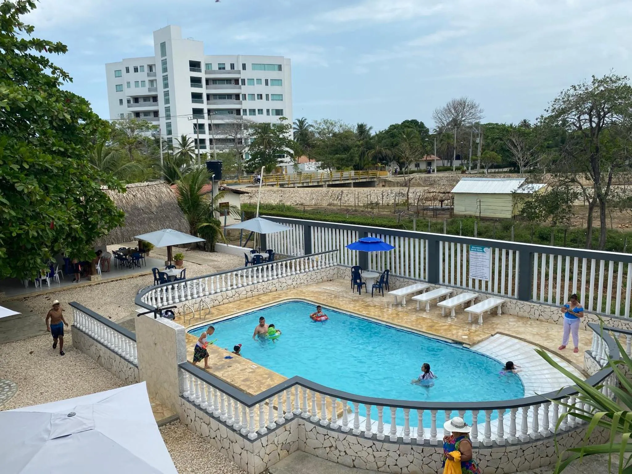 Une piscine rectangulaire avec des personnes s'amusant dans l'eau, entourée d'une terrasse et de bâtiments.
