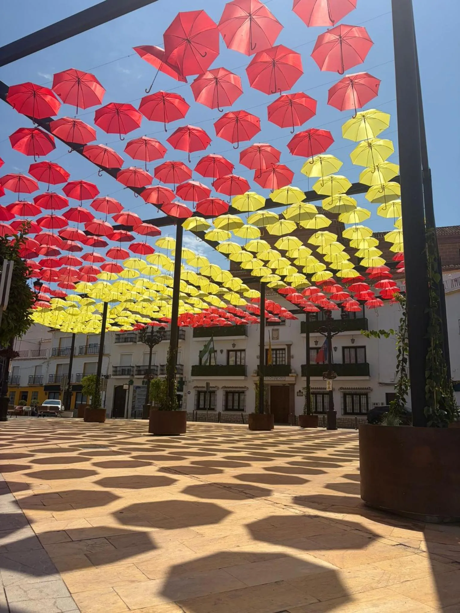 Une place piétonne est ombragée par une installation artistique de parasols colorés.