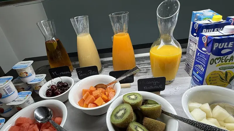 Une table présente des carafes de jus de fruits, des yaourts, des fruits frais coupés et des céréales.