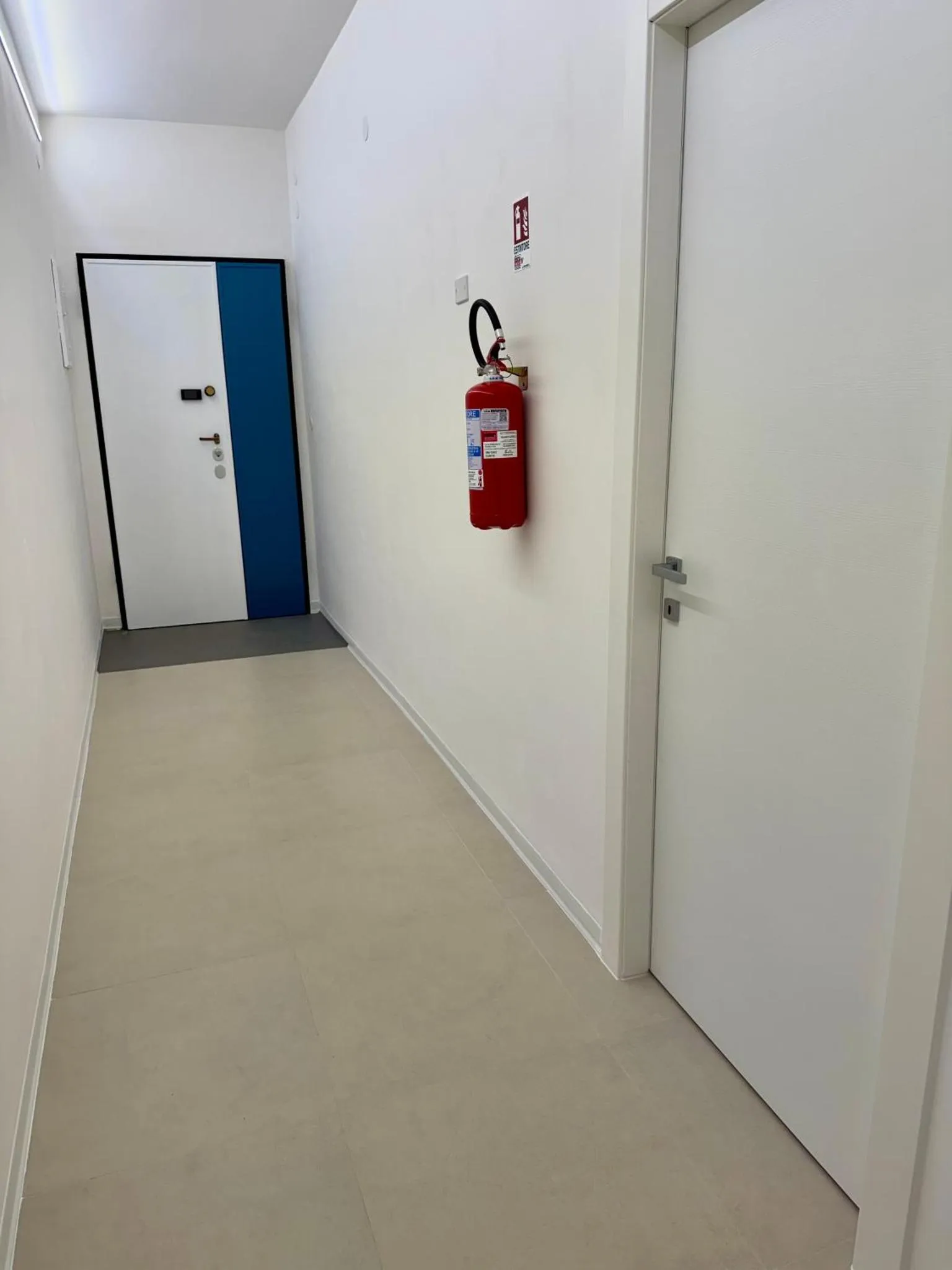 Un couloir d'hôtel blanc avec des portes et un extincteur rouge.