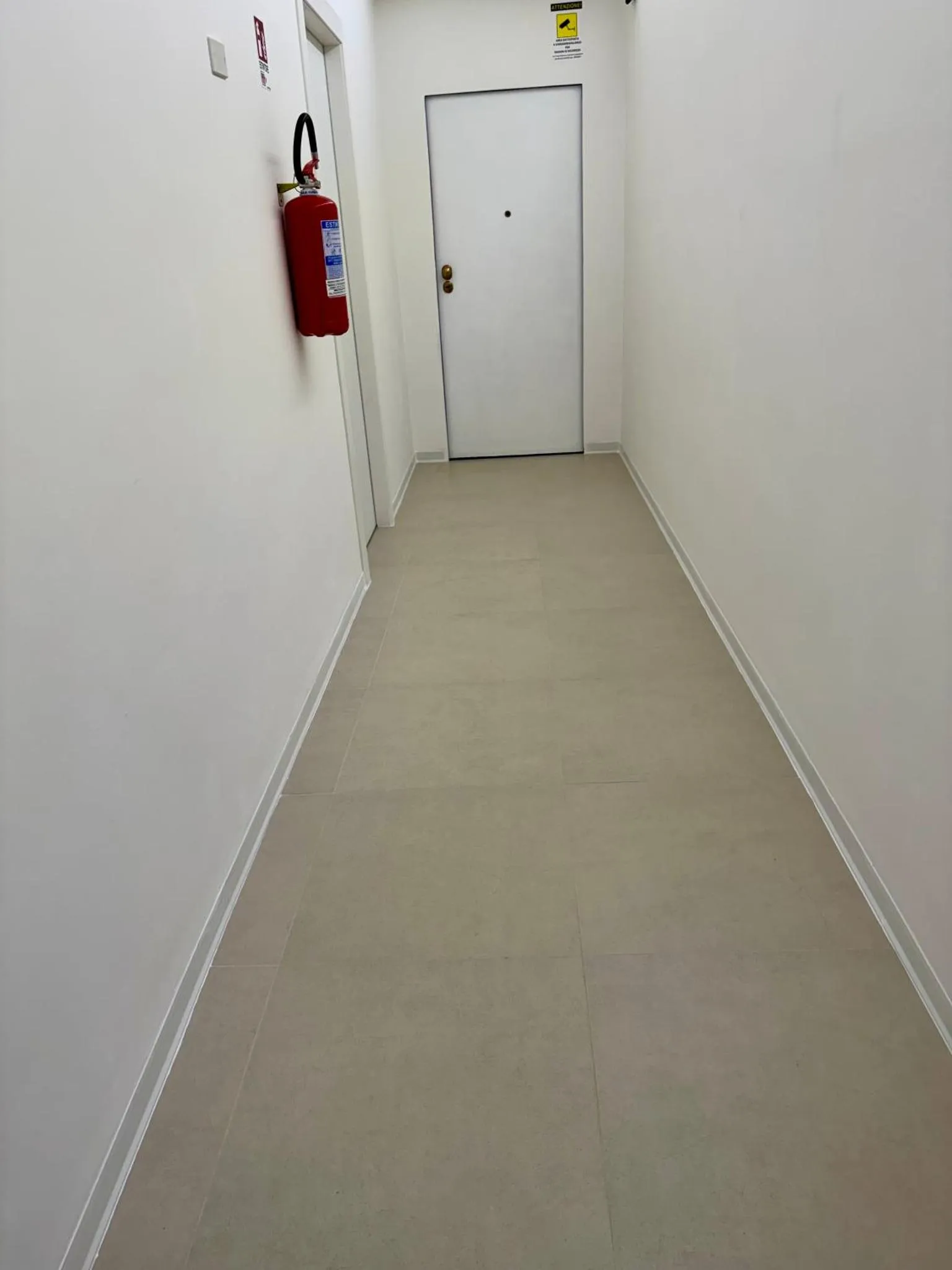 Un couloir intérieur aux murs clairs et au sol carrelé mène à une porte blanche.