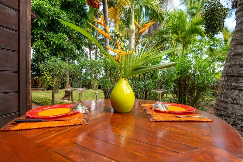 Une table ronde en bois est dressée pour un repas en extérieur, décorée de vaisselle colorée et de fleurs tropicales.