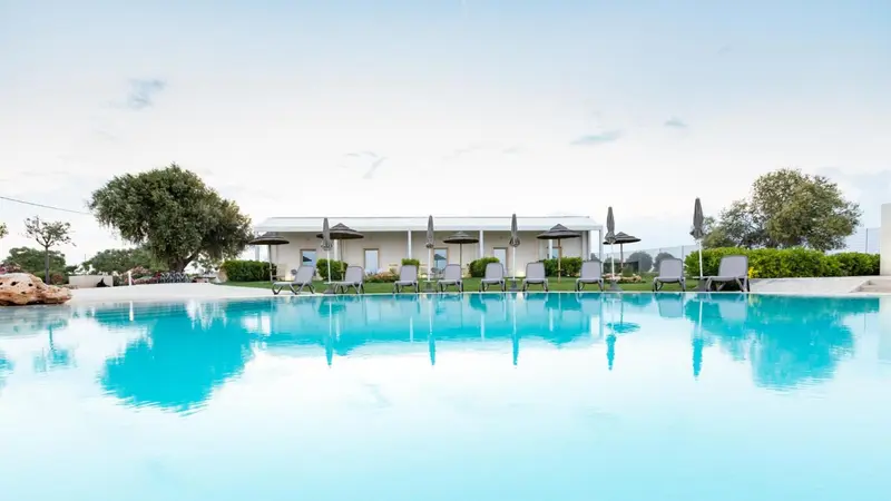 Une piscine turquoise reflète un bâtiment blanc avec des transats et des parasols.