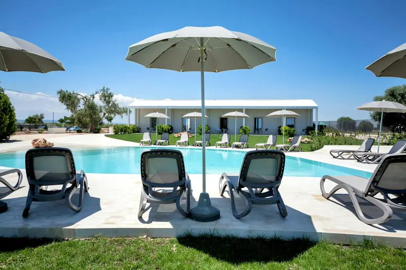 Transats et parasols bordent une piscine turquoise devant un bâtiment blanc.