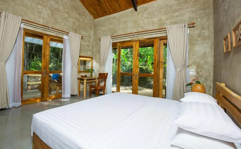 Une chambre lumineuse avec un grand lit et des baies vitrées ouvertes sur la végétation.
