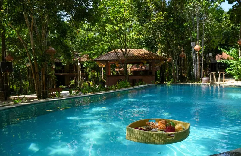 Une piscine turquoise reflète un bâtiment entouré de végétation luxuriante et un plateau de petit-déjeuner flotte à la surface.