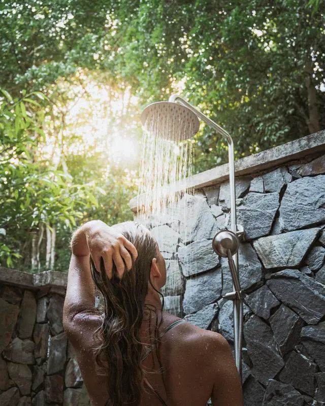 Une personne se rince les cheveux sous une douche extérieure en pierre.