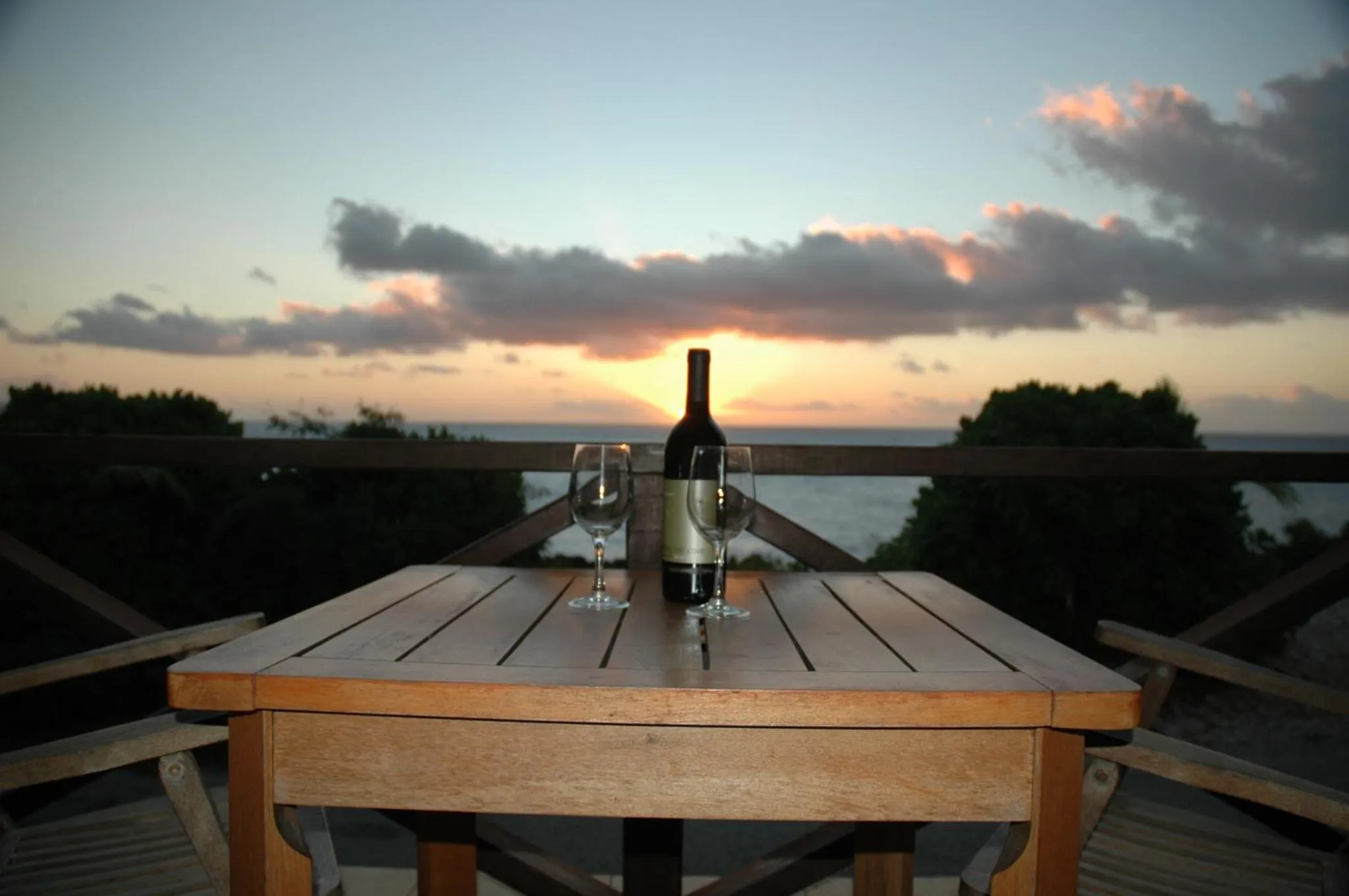 Une table en bois avec une bouteille de vin et deux verres, face à un coucher de soleil sur la mer.