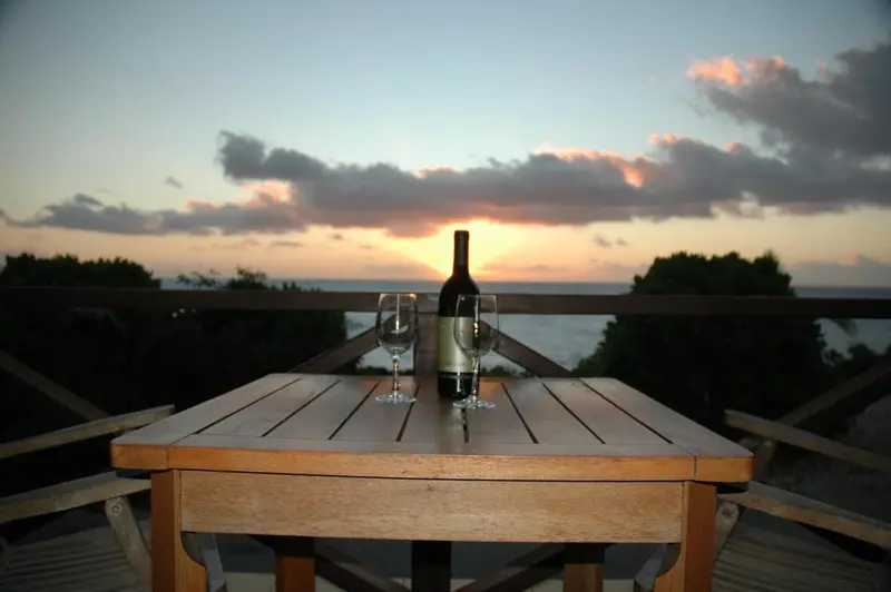 Une table en bois avec une bouteille de vin et deux verres, face à un coucher de soleil sur la mer.