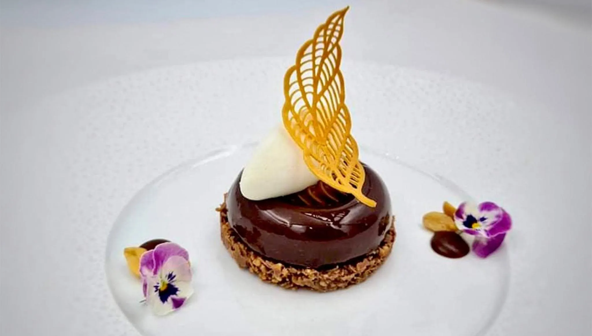 Une assiette blanche présente un dessert au chocolat décoré d'une feuille croustillante et de fleurs comestibles.