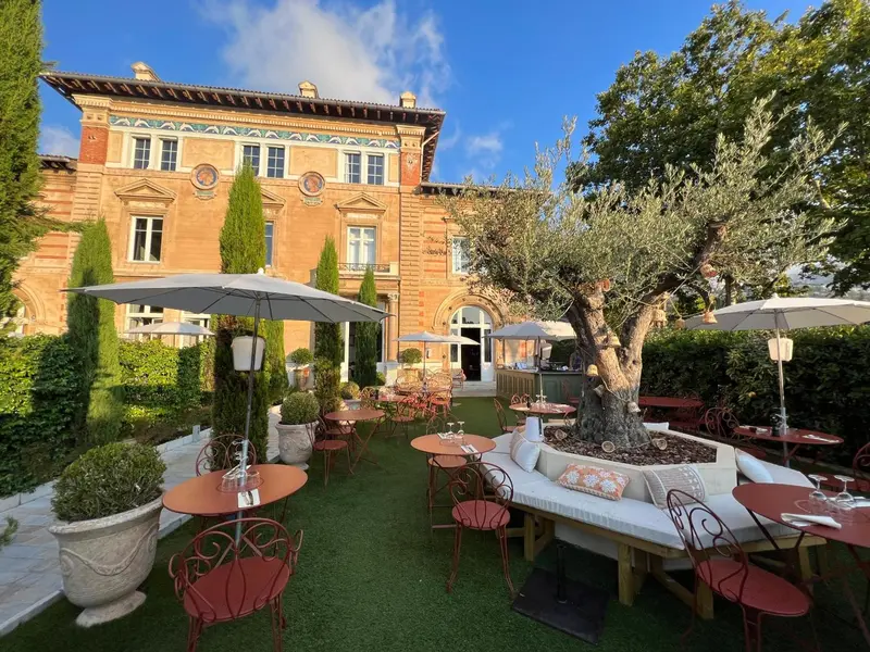 Un jardin verdoyant avec des tables et des chaises rouges devant un bâtiment en pierre.