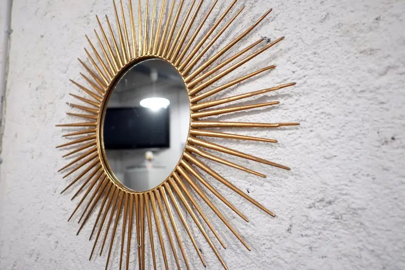 Miroir rond doré avec des rayons en métal sur un mur texturé.