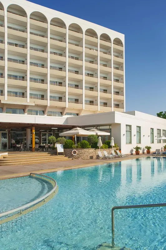 Bâtiment blanc à plusieurs étages surplombant une piscine turquoise et un espace aménagé avec des transats.