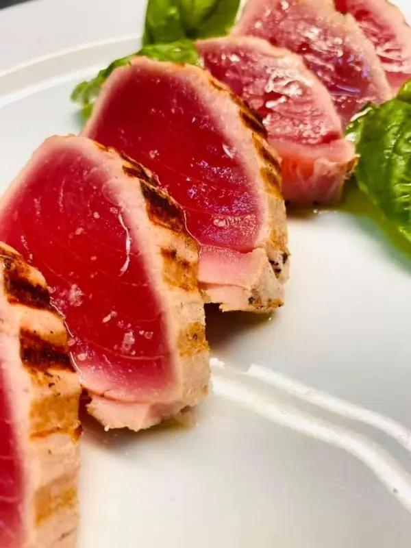 Tranches de thon rouge grillé sur une assiette blanche, garnies de feuilles vertes.