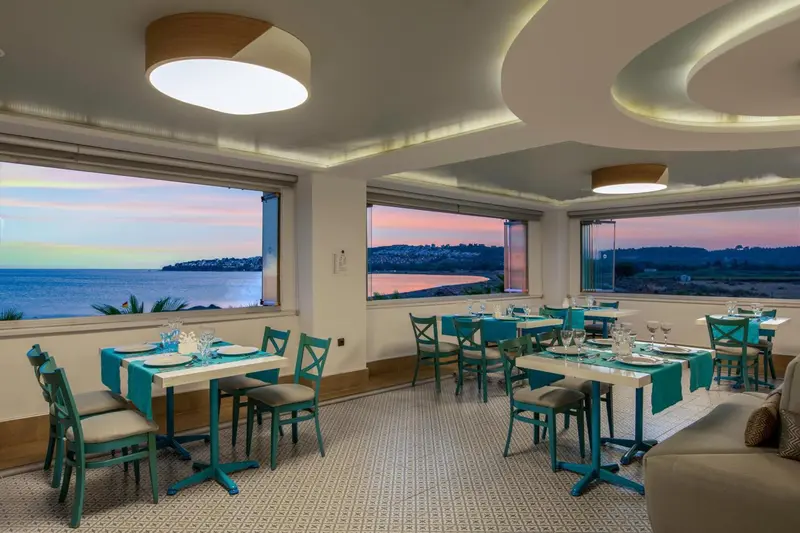 Une salle lumineuse avec des tables dressées face à une vue sur la mer au coucher du soleil.