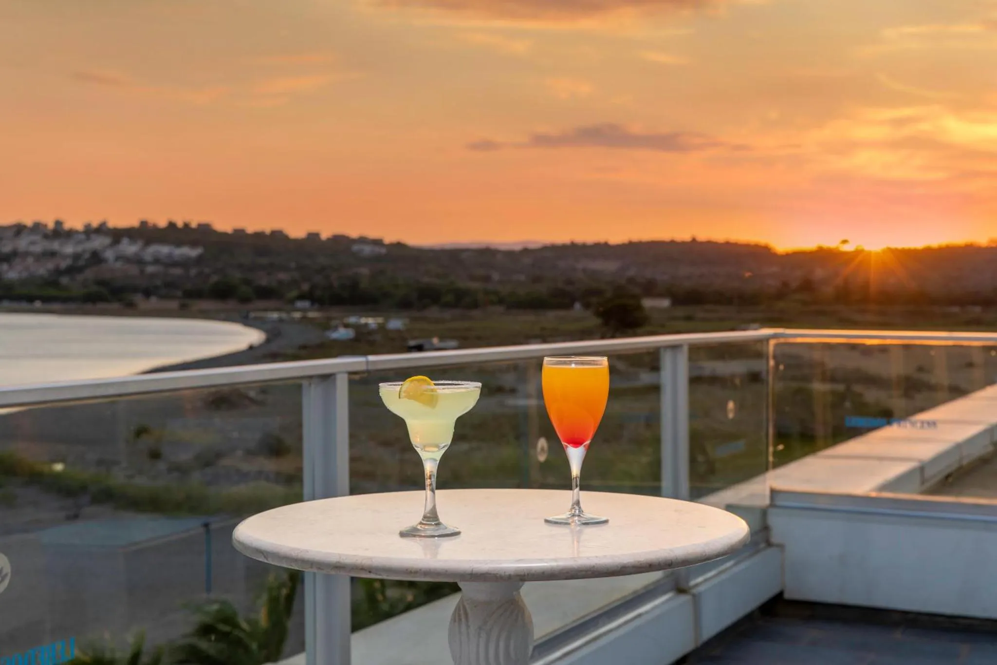 Deux cocktails colorés sur une table ronde en pierre, avec une vue sur la mer et un coucher de soleil.