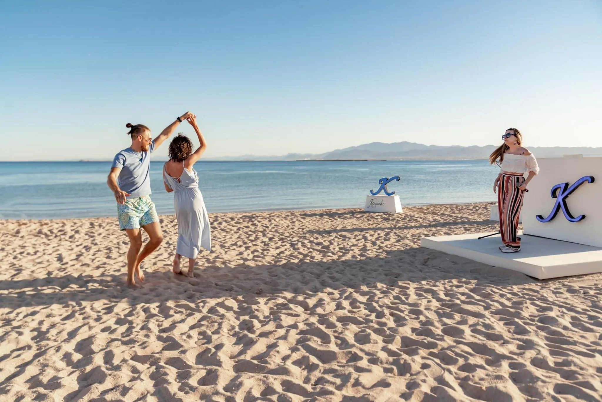 Deux personnes se photographient sur une plage de sable fin avec une vue sur la mer.