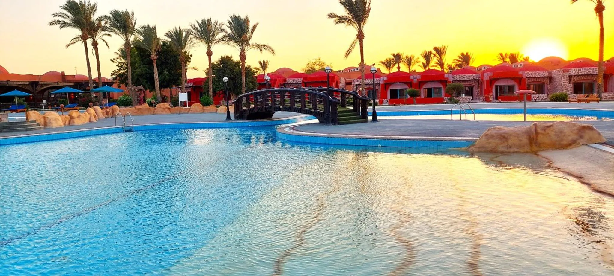 Une piscine turquoise au premier plan, avec des bâtiments rouges et des palmiers au loin, baignés par la lumière dorée du coucher de soleil.