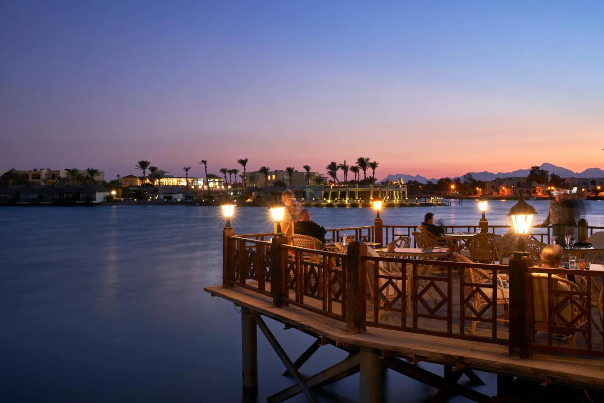 Une terrasse en bois avec des tables et des chaises au bord de l'eau au coucher du soleil.