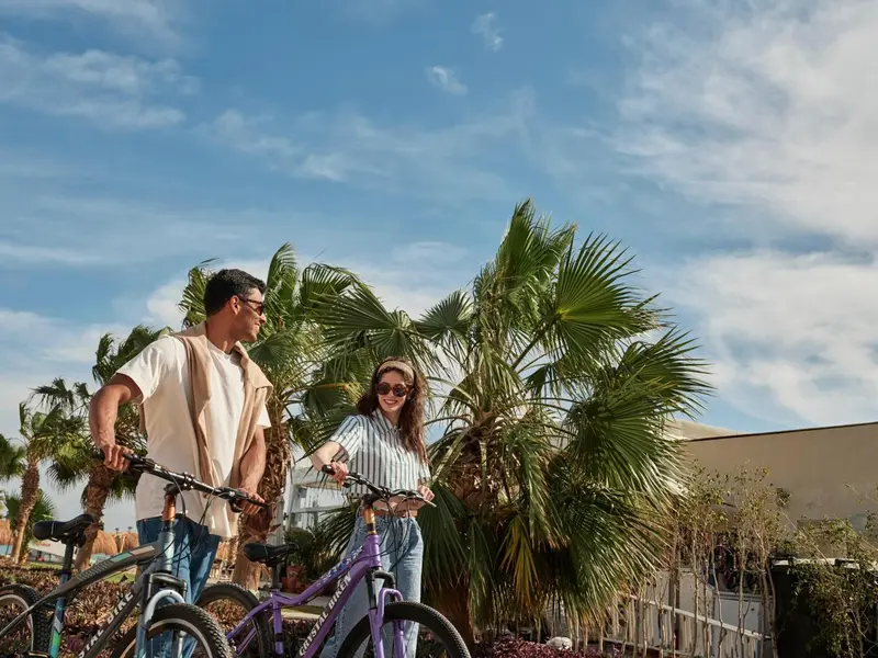Un couple se promène à vélo devant des palmiers et un bâtiment.