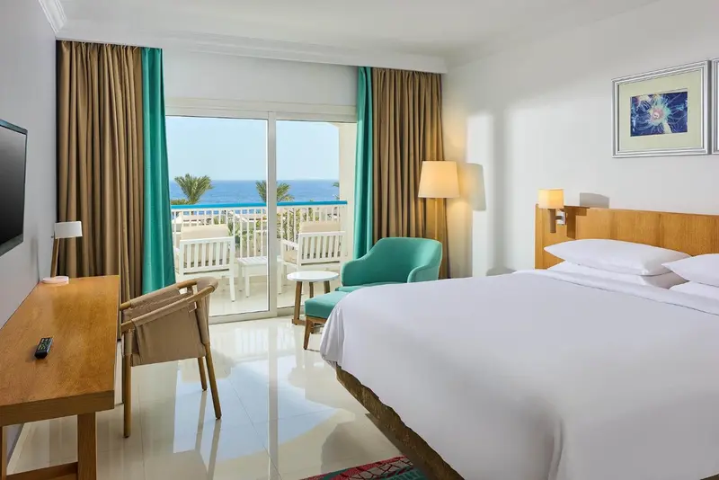 Chambre lumineuse avec un lit blanc et un accès à un balcon avec vue sur la mer.