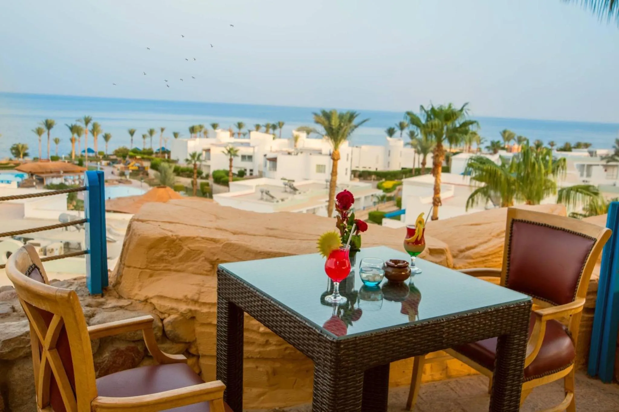 Une table basse avec des boissons colorées est mise en scène sur une terrasse ensoleillée avec vue sur la mer.