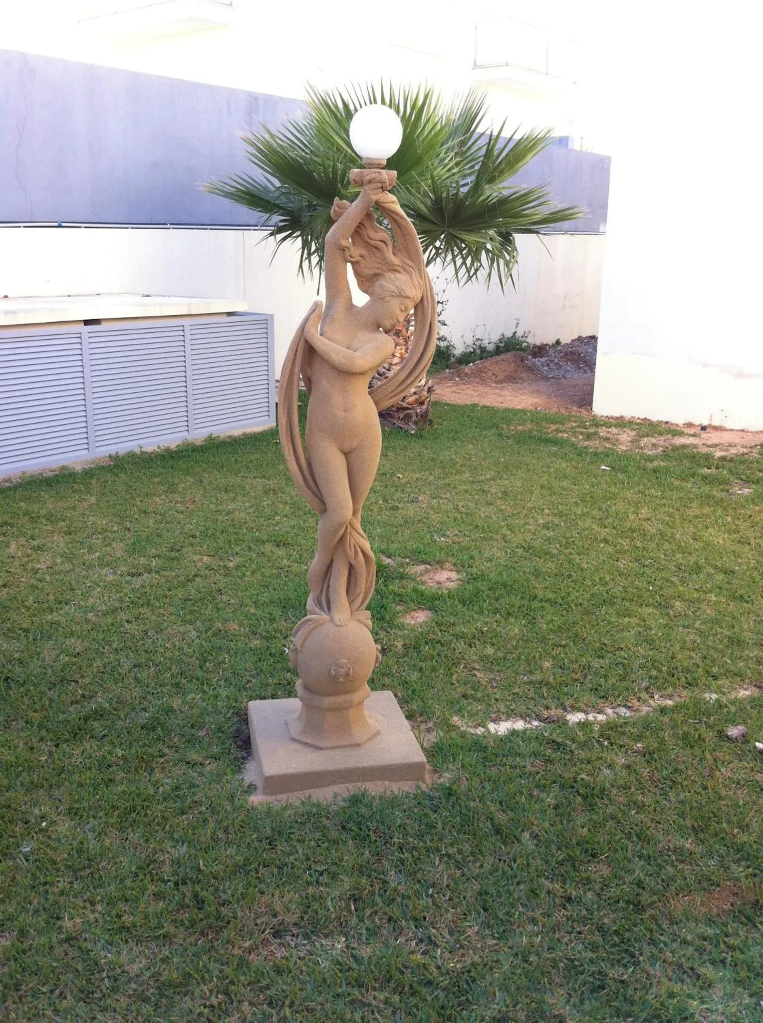 Statue de femme adossée à un palmier sur une pelouse.