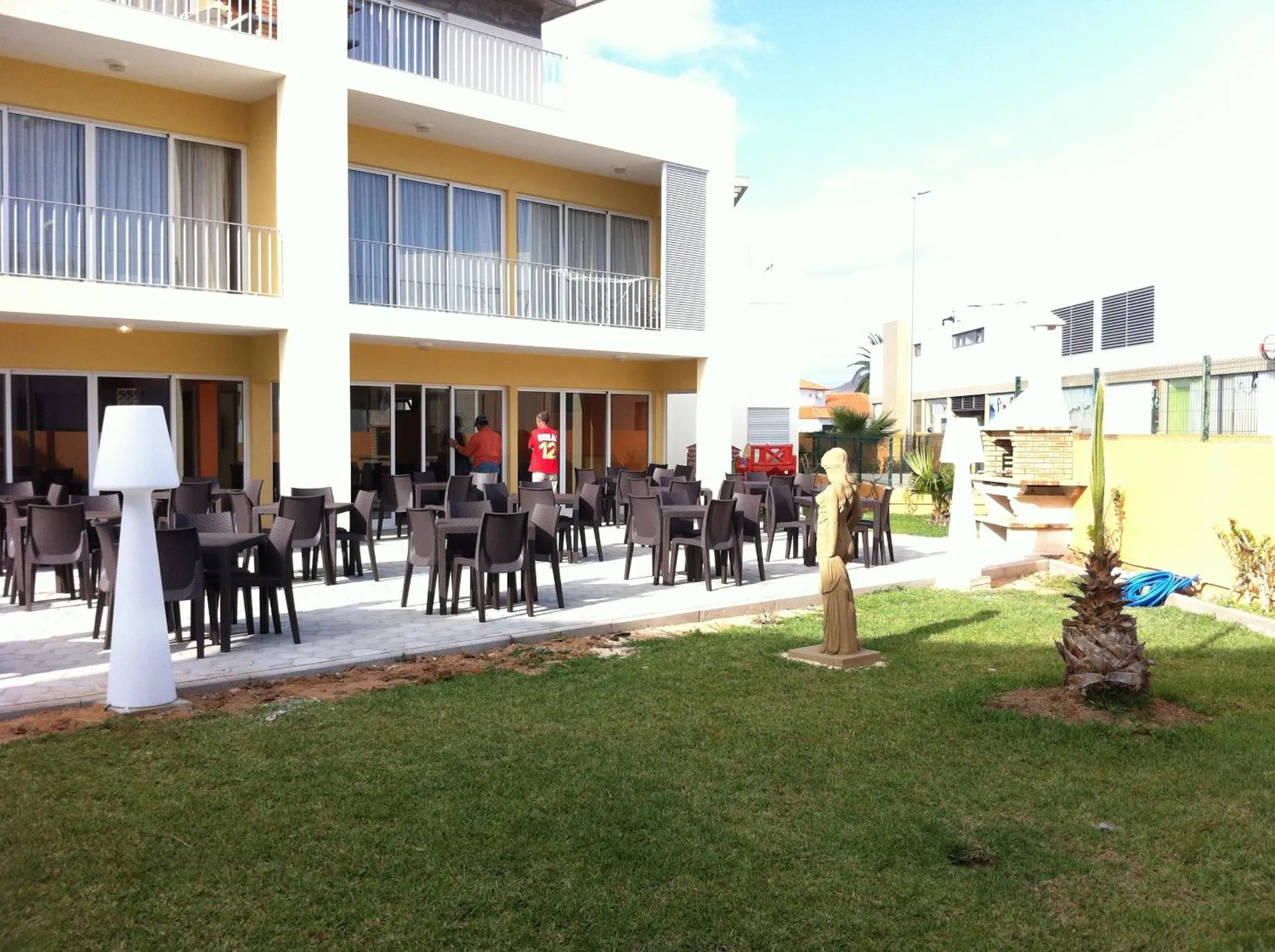 Terrasse ombragée avec tables et chaises, devant un bâtiment jaune et blanc.